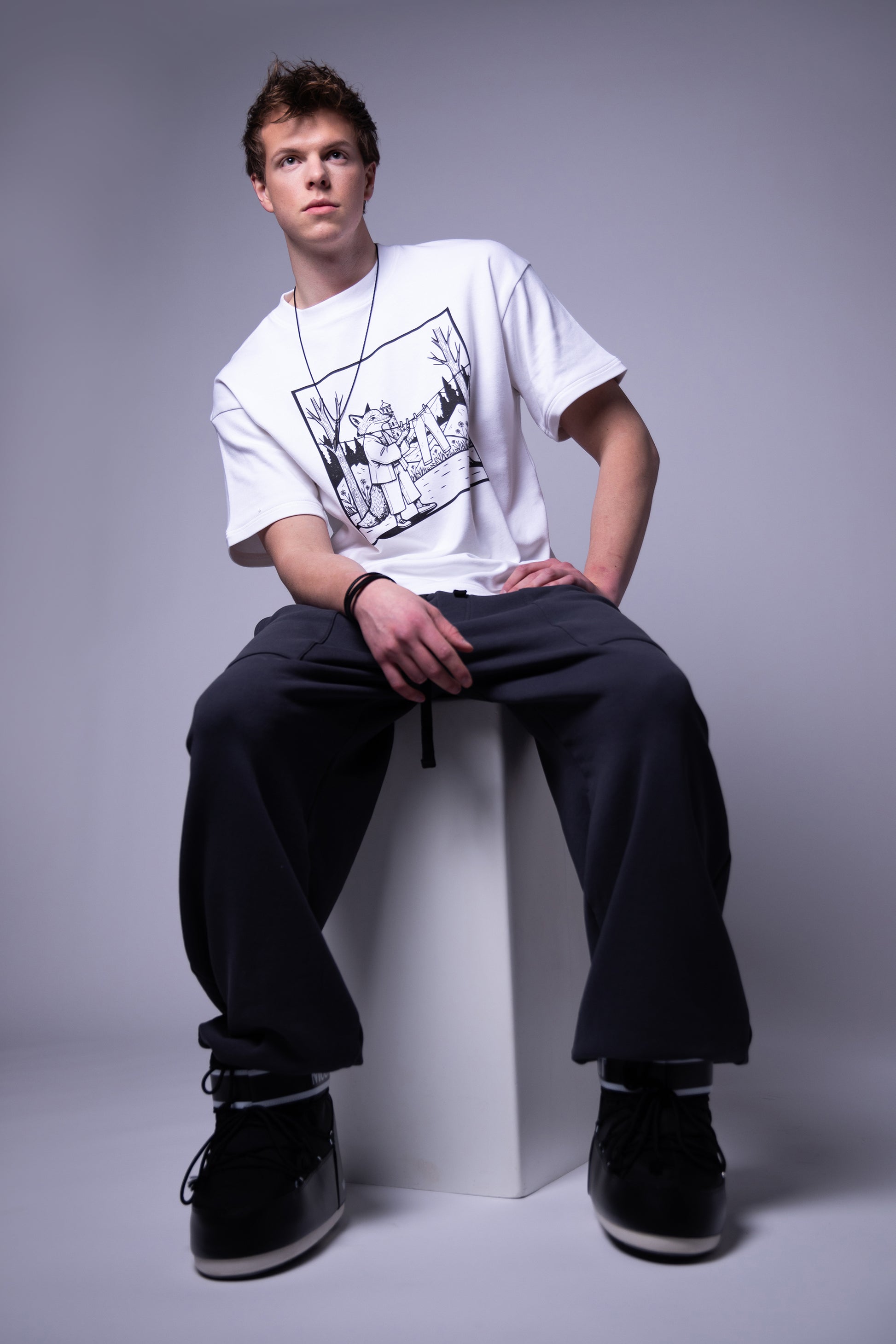 Model seated wearing white Supima cotton Lineage Tee with black graphic and deep grey sweatpants